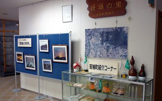 ボードに飾られてある5枚の絵画や、ガラスケースの中や上にひょうたん、本などが置かれてある斑鳩町紹介コーナーの写真