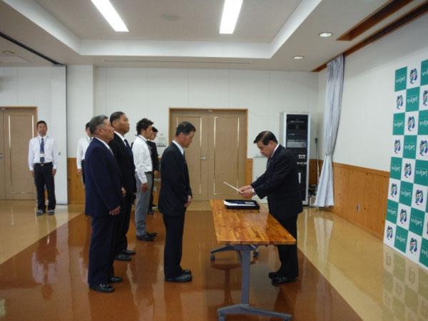 辞令を読み上げている町長と、町長の前に向かい合って立っている中村猟友会長、会長の後ろに横一列に整列して立っている4人の男性、部屋の隅に建っている2人の男性の写真