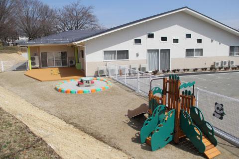 白い外壁で平屋づくりのいいっ子センターの建物と駐車場、庭にある遊具や砂場の写真