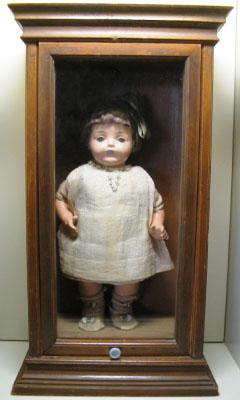木のフレームのガラスケースに入った、白い服を着ている青い目の人形の写真
