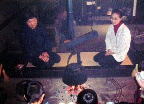 天井から吊り下げられたやかんが火に掛かっている囲炉裏の前に女性二人が正座をして座っている写真