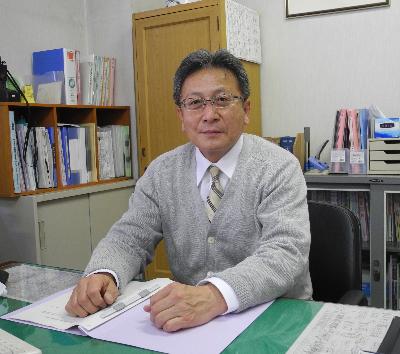 片桐教育長が書類の置かれた机に座りカメラ目線で写っている写真