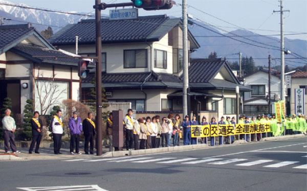 「春の交通安全運動実施中」と書かれた看板やのぼり旗を持って沿道に立っている警察官や住民の写真