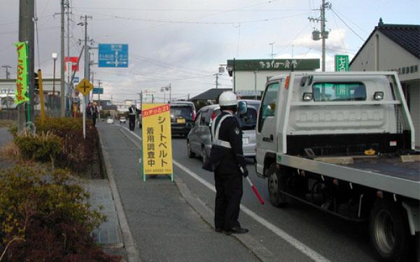 歩道に「シートベルト着用調査中」の看板が立てられており、その付近で警察官が通行する車をチェックしている写真