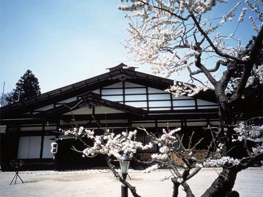 平屋づくりの古い日本家屋風の飯島陣屋と、白い花が咲いた梅の木の写真